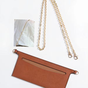 Könnte beinhalten: Eine braune Leder-Handyhülle mit goldener Kette. Die Hülle hat eine rechteckige Form mit einem Ausschnitt für das Telefon und zwei kleine goldene Ringe an den Seiten. Die goldene Kette hat an jedem Ende einen Karabinerverschluss.