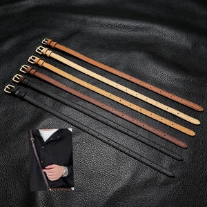 May include: Five thin leather straps in varying shades of brown and black, each featuring a gold-toned metal buckle and multiple adjustment holes. The straps are arranged diagonally, showcasing a smooth finish and consistent stitching along the edges.