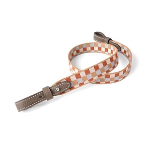 May include: A woven camera strap with a geometric pattern in shades of brown, orange, and blue. The strap has leather ends in a matching brown color with white stitching and metal hardware.