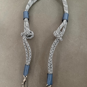 Op de afbeelding: Een zwart-wit patroon touwriem met blauwe lederen accenten en zilverkleurige sluitingen. De riem is bovenaan geknoopt en heeft een herhalend geometrisch ontwerp. De riem is ontworpen voor een tas of portemonnee.