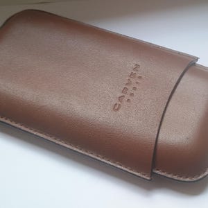 Può includere: Un astuccio in pelle marrone con il marchio in rilievo "CARVEN PARIS". L'astuccio ha una forma rettangolare con angoli arrotondati e una chiusura a patta. Le cuciture sono visibili sui bordi.