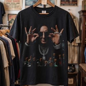 May include: A black short-sleeve t-shirt featuring a graphic print of a person wearing round dark sunglasses, multiple silver chains, and rings on their fingers. The person is positioned behind a collection of small glass bottles.