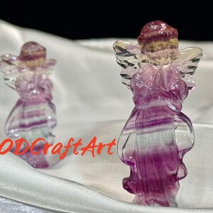 Puede incluir: Dos figuras de ángeles talladas en cristal de fluorita. Los ángeles están de pie con alas y túnicas fluidas. El cristal es una mezcla de morado, rosa, amarillo y transparente. El texto "RODCraftArt" está en la esquina inferior izquierda.