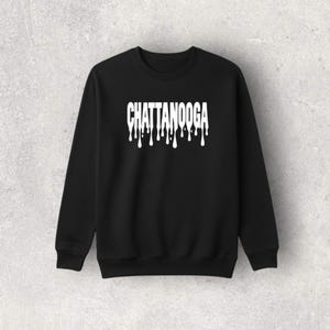 May include: Black sweatshirt with the word "CHATTANOOGA" in white, dripping font across the chest. The long-sleeved crewneck top is a solid black color.