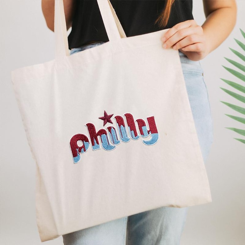 May include: Cream-colored tote bag with the embroidered word "Philly" in red and blue. The word is stylized with a star above the "i". The bag has two long handles and is held by a person.