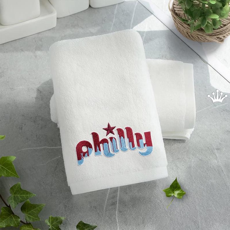 May include: White bath towel with the word "Philly" embroidered in red and blue, with a star accent. The towel is folded and stacked with another white towel. The background is a light gray surface.