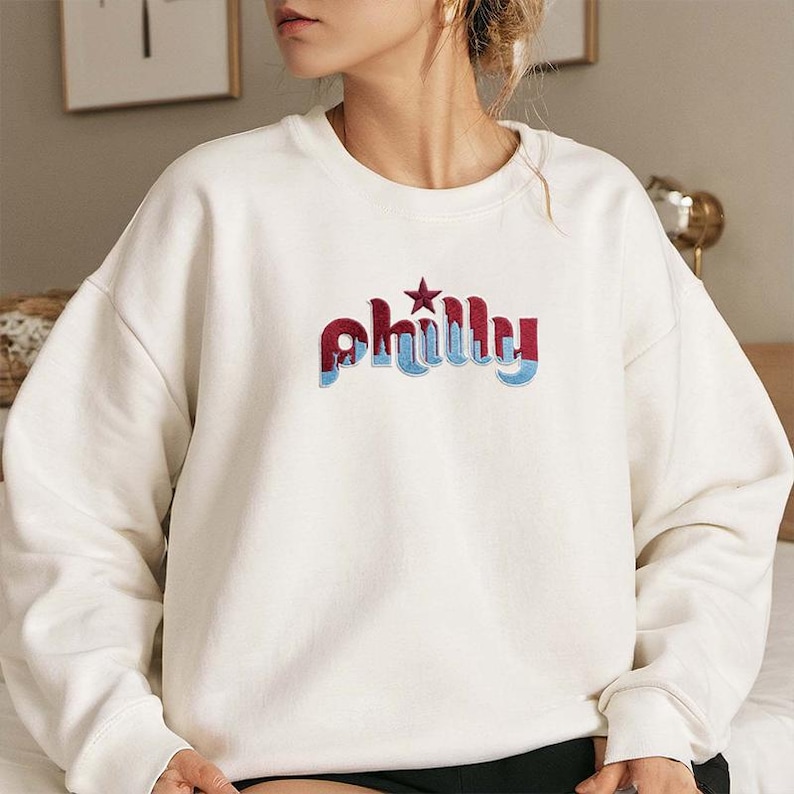 May include: Cream-colored sweatshirt with the word "Philly" embroidered across the chest in red, blue, and white. The word is stylized with a star above the "i". The sweatshirt has a crew neck and long sleeves.