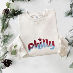 May include: A cream-colored crewneck sweatshirt featuring the word "philly" embroidered in a two-tone design with dark red on top and light blue on the bottom. A dark red star is embroidered above the letter i in the word philly.