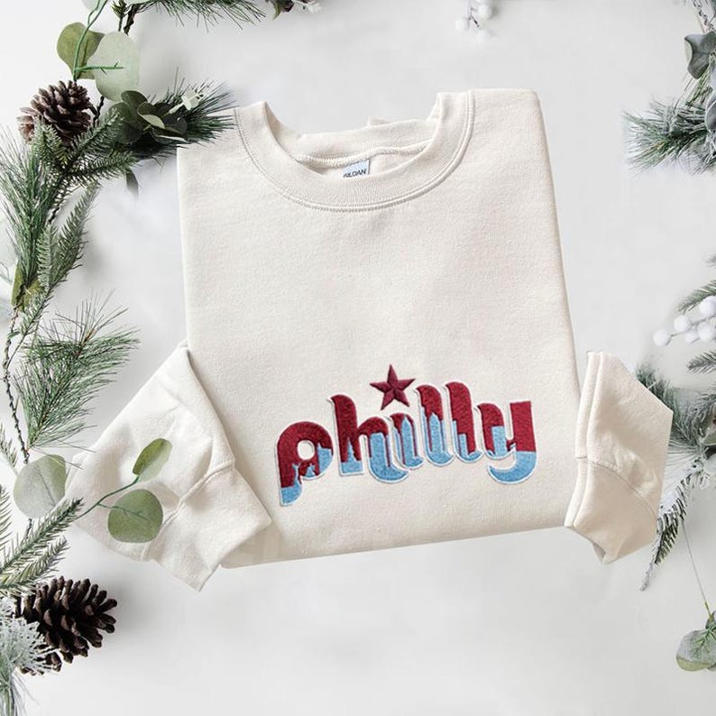 May include: A cream-colored crewneck sweatshirt featuring the word "philly" embroidered in a two-tone design with dark red on top and light blue on the bottom. A dark red star is embroidered above the letter i in the word philly.