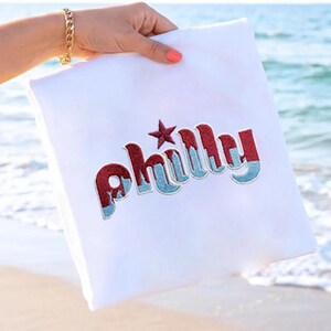 May include: White square pillow with the word "Philly" embroidered in red and blue, with a red star above the "h". The pillow is held by a person on a beach with the ocean in the background.