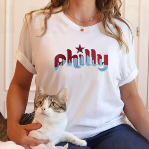 May include: White t-shirt with rolled sleeves featuring the word "Philly" in maroon and light blue, with a star above the "y". A cat is in the foreground. The shirt is worn by a person.