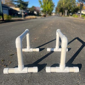May include: Two white PVC pipe parallette bars are positioned on a paved surface. Each bar consists of a horizontal base, vertical supports, and a top bar. The bars are designed for calisthenics and bodyweight training.