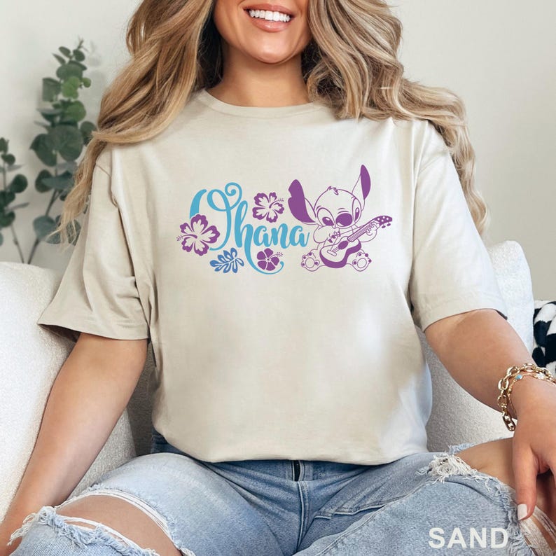May include: Sand-colored t-shirt featuring the word "Ohana" in blue and purple script, accompanied by floral designs and a purple cartoon character playing a guitar. The shirt is made of a soft material.