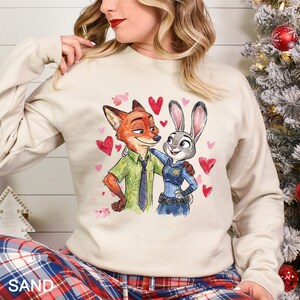May include: A sand-colored sweatshirt featuring a watercolor illustration of Nick Wilde and Judy Hopps from Zootopia, surrounded by pink and red hearts. Nick is wearing a green shirt and tie, while Judy is in a police uniform.