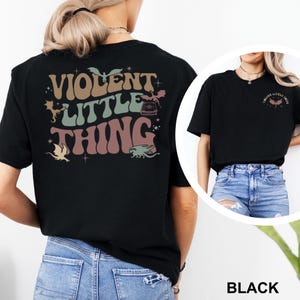 May include: Black t-shirt with the text "VIOLENT LITTLE THING" in a retro font, with dragon and star graphics. The shirt is shown on a person wearing blue jeans. A smaller version of the design is on the front of the shirt in the inset image.