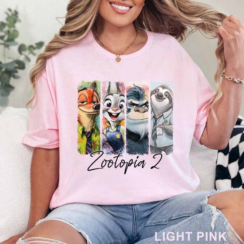 May include: Pale pink t-shirt with a vibrant graphic of Zootopia characters. The design showcases Nick Wilde, Judy Hopps, a gorilla, and Flash the sloth, with "Zootopia 2" written underneath. The shirt is made of a soft material.