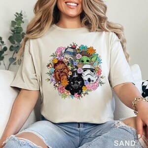 May include: Sand-colored t-shirt featuring a floral wreath design with Star Wars characters including Chewbacca, Darth Vader, a Stormtrooper, R2-D2, and Grogu. The shirt has short sleeves and a crew neck.