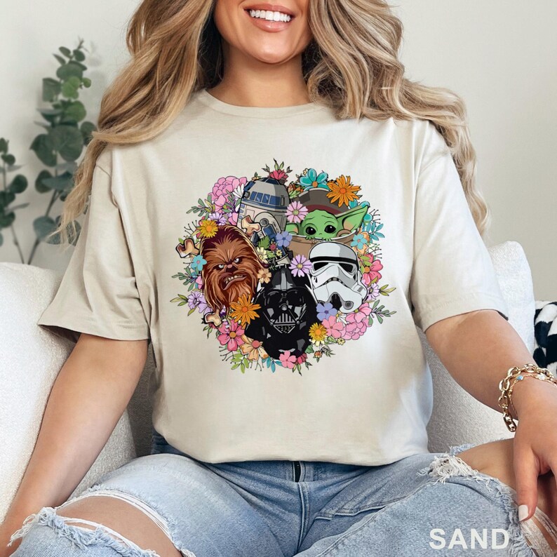 May include: Sand-colored t-shirt featuring a floral wreath design with Star Wars characters including Chewbacca, Darth Vader, a Stormtrooper, R2-D2, and Grogu. The shirt has short sleeves and a crew neck.