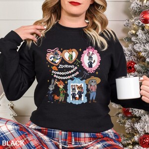 Op de afbeelding: Zwarte crewneck sweatshirt met een kleurrijke afbeelding van personages uit de animatiefilm Zootopia. Het ontwerp bevat ingelijste portretten en hartvormige elementen met het woord "Zootopia". De sweatshirt wordt door een persoon gedragen.