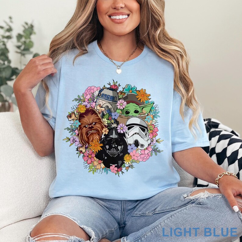 May include: Light blue t-shirt featuring a floral arrangement with Star Wars characters including Chewbacca, Darth Vader, a Stormtrooper, R2-D2, and Grogu. The text "LIGHT BLUE" is at the bottom.