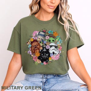 May include: Military green t-shirt featuring a floral design with Star Wars characters including Darth Vader, Chewbacca, and Baby Yoda. The shirt has a crew neck and short sleeves. The text "MILITARY GREEN" is at the bottom.