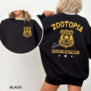 May include: Black sweatshirt featuring a gold Zootopia Police Department badge on the front and back. The back design includes the text "Zootopia Police Department" and "Now Recruiting!" in gold. The sweatshirt has a white collar.
