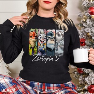 Op de afbeelding: Zwarte sweatshirt met een kleurrijke afbeelding van personages uit "Zootopia 2". Het ontwerp bevat Nick Wilde, Judy Hopps en andere personages. De woorden "Zootopia 2" zijn in cursieve letters geschreven. Het woord "BLACK" staat onderaan gedrukt.