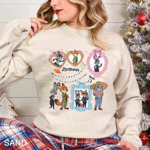 Op de afbeelding: Zandkleurige sweatshirt met stripfiguren uit de film Zootopia. Het ontwerp bevat ingelijste afbeeldingen van de personages, het woord "Zootopia" en de zin "welcome to zootopia". De sweatshirt heeft lange mouwen.