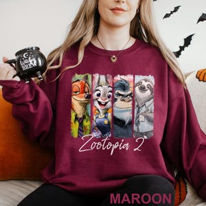 Op de afbeelding: Bordeaux sweatshirt met personages uit Zootopia 2. Het ontwerp toont Nick Wilde, Judy Hopps, Chief Bogo en Flash. De tekst "Zootopia 2" is in witte letters geschreven. Het woord "MAROON" staat onderaan.