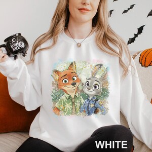 Puede incluir: Sudadera blanca con un gr&aacute;fico colorido de Nick Wilde y Judy Hopps de Zootopia. El dise&ntilde;o muestra a los personajes con sus respectivos atuendos. La palabra "WHITE" est&aacute; impresa en la parte inferior de la sudadera.
