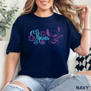 May include: Navy blue t-shirt featuring the word "Ohana" in blue and purple floral design, with a purple cartoon character playing a guitar. The shirt is made of a soft material.