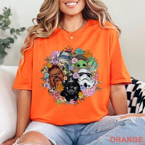 May include: Orange t-shirt featuring a floral arrangement with Star Wars characters including Chewbacca, Darth Vader, a Stormtrooper, R2-D2, and Grogu. The word "ORANGE" is printed at the bottom. The shirt is made of a soft material.