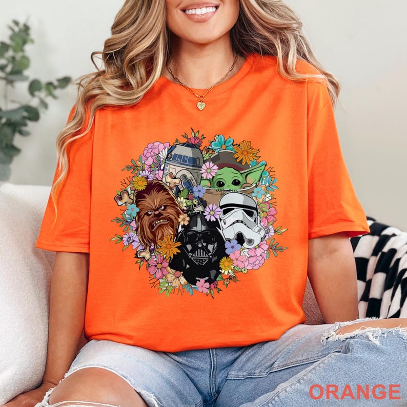 May include: Orange t-shirt featuring a floral arrangement with Star Wars characters including Chewbacca, Darth Vader, a Stormtrooper, R2-D2, and Grogu. The word "ORANGE" is printed at the bottom. The shirt is made of a soft material.
