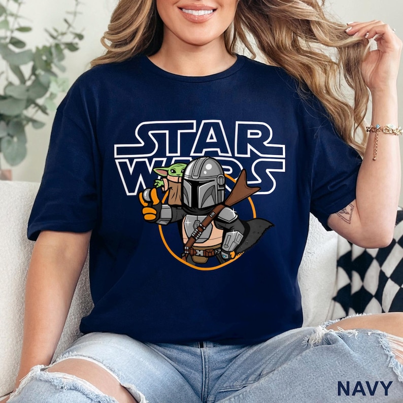 May include: Navy blue t-shirt featuring a cartoon illustration of The Mandalorian and Grogu from Star Wars. The Star Wars logo is above the characters. The word "NAVY" is printed at the bottom right corner of the shirt.