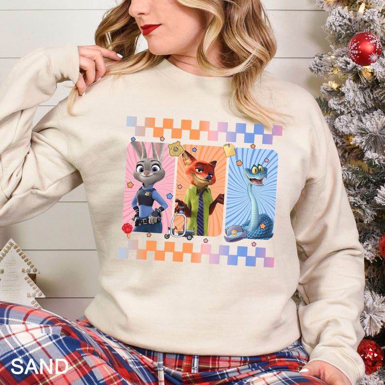 May include: A sand-coloured sweatshirt featuring three cartoon characters: a rabbit, a fox, and a snake. The characters are set against colourful backgrounds with a checkered pattern. The sweatshirt is a comfortable, casual style.