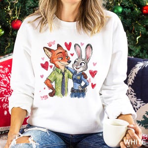 May include: White sweatshirt featuring a watercolor illustration of Nick Wilde and Judy Hopps from Zootopia, surrounded by red hearts. Nick is a fox in a green shirt and tie, and Judy is a rabbit in a police uniform.