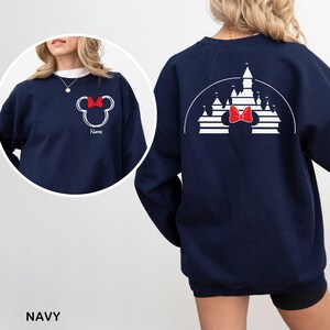 Pu&ograve; includere: Felpa blu navy con un design di Minnie Mouse bianco e un fiocco rosso, con la parola "Name". Il retro presenta un design a castello bianco e un fiocco rosso. La parola "NAVY" &egrave; in basso nell'immagine.