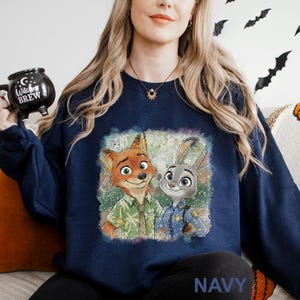 Puede incluir: Sudadera azul marino con un gr&aacute;fico colorido de Nick Wilde y Judy Hopps de Zootopia. La imagen muestra a los personajes en un estilo art&iacute;stico y vibrante. La palabra "NAVY" est&aacute; impresa en la parte inferior de la sudadera.