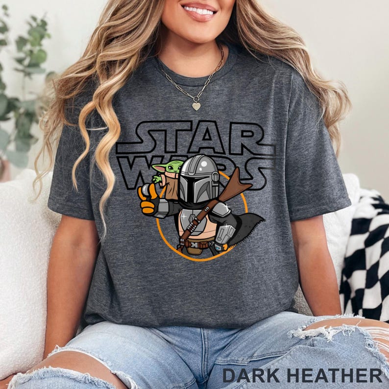 May include: Dark heather gray t-shirt featuring a cartoon illustration of The Mandalorian and Grogu from Star Wars. The Star Wars logo is above the illustration. The text "DARK HEATHER" is at the bottom of the shirt.