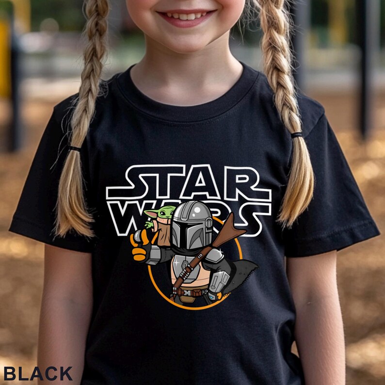 May include: Black t-shirt featuring the Star Wars logo and a cartoon illustration of The Mandalorian and Grogu. The design includes a silver helmet, brown rifle, and a yellow circle. The word "BLACK" is printed at the bottom.