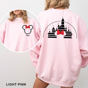 Pu&ograve; includere: Felpa rosa chiaro con fiocco di Minnie Mouse e nome personalizzato sul davanti. Il retro presenta una silhouette di castello nero e un fiocco rosso. Il testo "LIGHT PINK" &egrave; in basso.