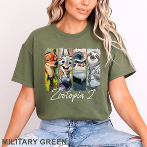 May include: Military green t-shirt with a vibrant graphic of Zootopia characters: Nick Wilde, Judy Hopps, Chief Bogo, and Flash. The text "Zootopia 2" is written in a cursive font below the characters. A casual, comfortable tee.