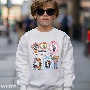 Op de afbeelding: Witte sweatshirt met kleurrijke cartoonfiguren en het woord "Zootopia". Het ontwerp bevat ingelijste afbeeldingen van dieren en de zin "welcome to Zootopia". De sweatshirt wordt gedragen door een kind met een zonnebril.