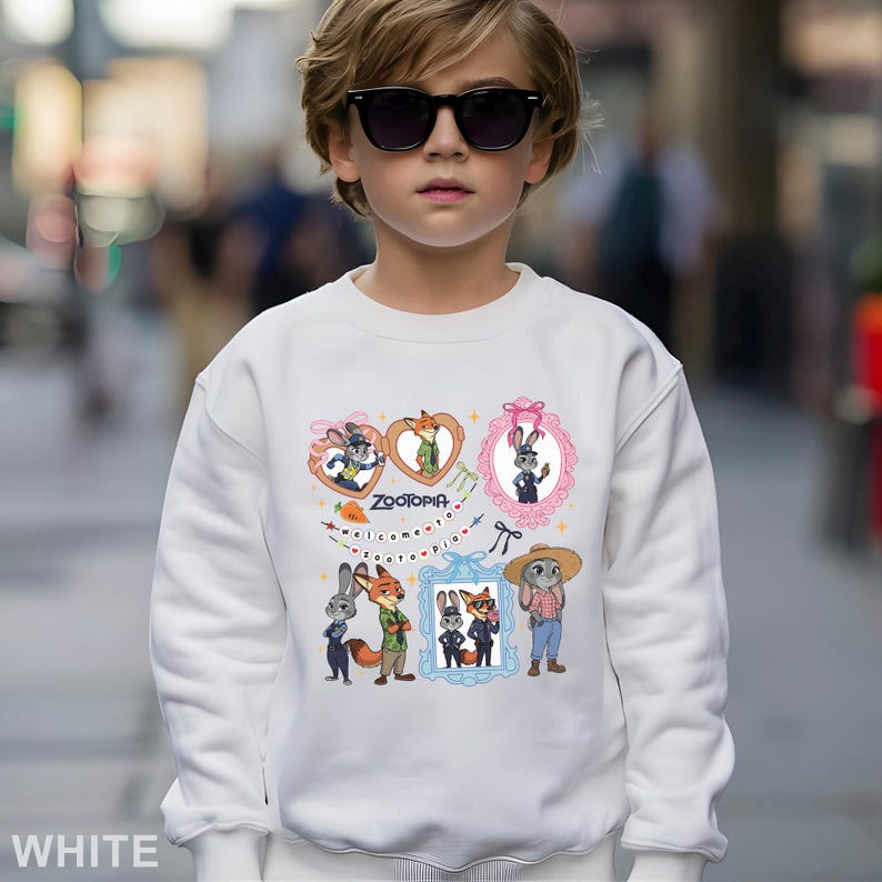 Op de afbeelding: Witte sweatshirt met kleurrijke cartoonfiguren en het woord "Zootopia". Het ontwerp bevat ingelijste afbeeldingen van dieren en de zin "welcome to Zootopia". De sweatshirt wordt gedragen door een kind met een zonnebril.