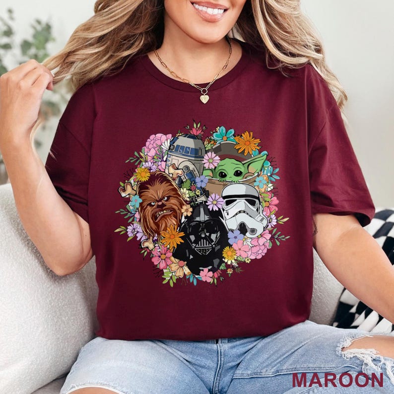 May include: Maroon t-shirt featuring a floral wreath design with Star Wars characters including Chewbacca, Darth Vader, a Stormtrooper, R2-D2, and Grogu. The word "MAROON" is printed at the bottom right.