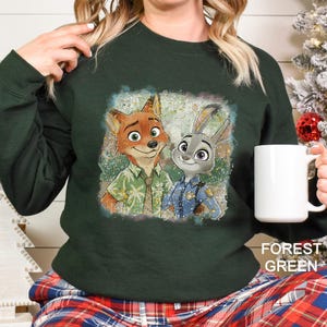 Puede incluir: Sudadera verde bosque con un gr&aacute;fico desgastado de Nick Wilde y Judy Hopps de Zootopia. La imagen muestra a los personajes con sus respectivos atuendos. El texto "FOREST GREEN" est&aacute; impreso en la parte inferior derecha.