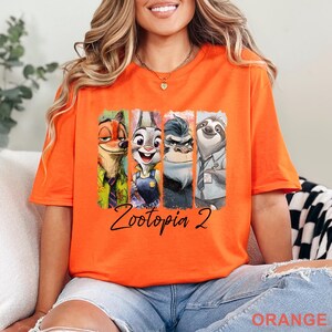 May include: Bright orange t-shirt with a graphic of characters from "Zootopia 2". The design shows Nick Wilde, Judy Hopps, Chief Bogo, and Flash. The words "Zootopia 2" are written in a cursive font below the characters.