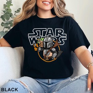 May include: Black t-shirt featuring a cartoon illustration of The Mandalorian and Grogu from Star Wars. The Star Wars logo is above the illustration. The word "BLACK" is printed at the bottom of the shirt.