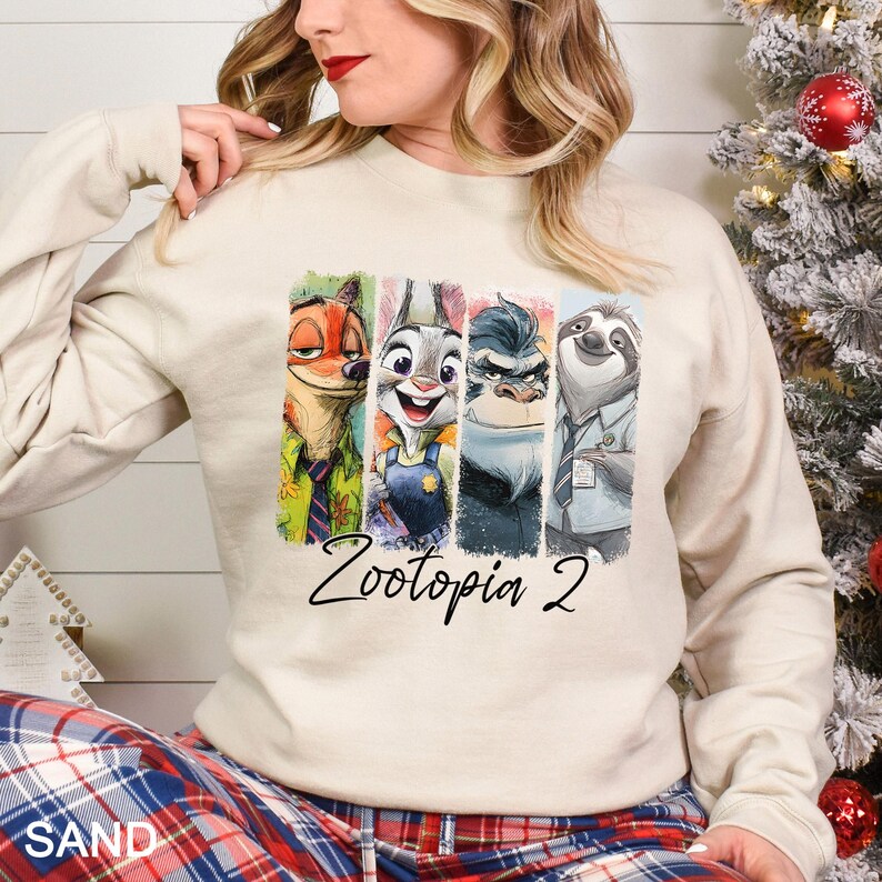 Op de afbeelding: Zandkleurige sweatshirt met een kleurrijke afbeelding van Zootopia-personages. De afbeelding bevat Nick Wilde, Judy Hopps, Chief Bogo en Flash. De tekst "Zootopia 2" staat eronder in een sierlijk lettertype.
