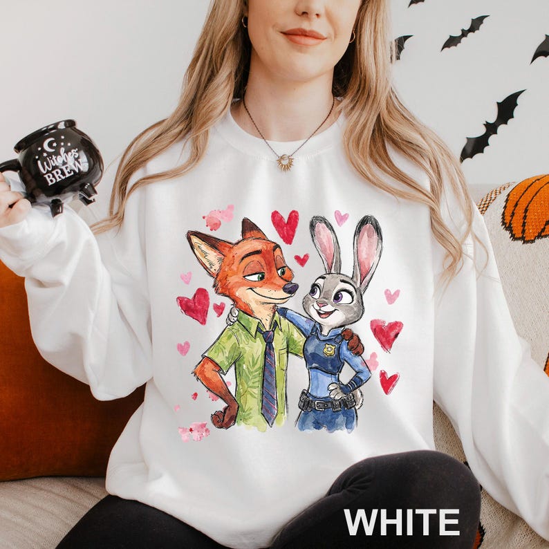 May include: White sweatshirt featuring a colorful illustration of Nick Wilde and Judy Hopps from Zootopia, surrounded by red hearts. The word "WHITE" is printed at the bottom. A black mug with the words "Witches Brew" is held in the upper left.